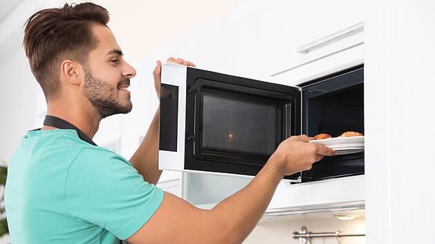 Junger Mann, der zu Hause einen Teller mit Croissants in den Mikrowellenherd stellt