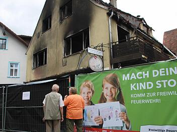 Passanten betrachten die ausgebrannte Asylunterkunft. Das einsturzgefährdete Haus ist mit Zäunen abgeriegelt, die von der Feuerwehr nach erfolgreich getaner Arbeit als Werbeflächen genutzt werden.Foto: Ekkehard Roepert