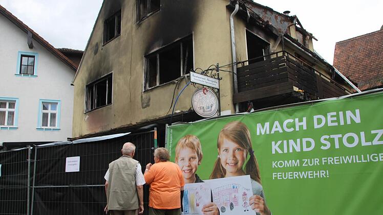 Passanten betrachten die ausgebrannte Asylunterkunft. Das einsturzgefährdete Haus ist mit Zäunen abgeriegelt, die von der Feuerwehr nach erfolgreich getaner Arbeit als Werbeflächen genutzt werden.Foto: Ekkehard Roepert