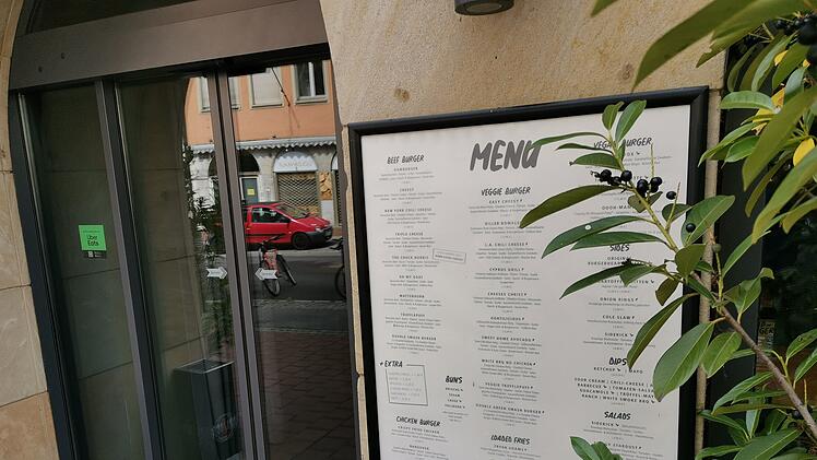 Ein Restaurant-Menü an einer Wand, daneben eine Eingangstür mit einem "Uber Eats"-Aufkleber. Im Vordergrund sind Pflanzen, im Hintergrund sieht man eine Straße mit Autos.