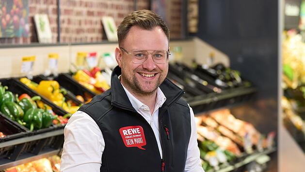Ein l&auml;chelnder Mann im REWE-Markt, umgeben von Obst- und Gem&uuml;seregalen, h&auml;lt ein Ger&auml;t in der Hand. Eine &Uuml;bernahme und Verbesserungen der Filiale in Baunach stehen bevor.