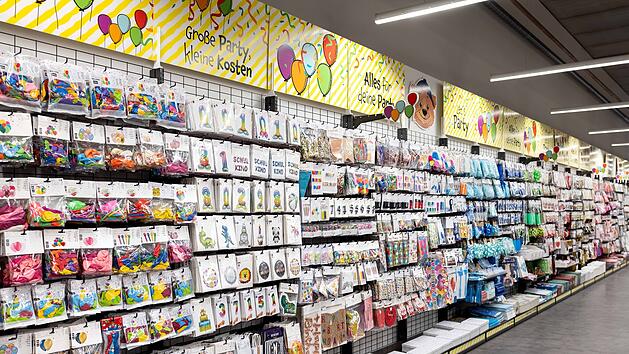 Ein gut sortiertes Ladenregal voller Partyartikel wie Ballons und Dekorationen. Die Beschriftung oben lautet: "Gro&szlig;e Party, kleine Kosten" und "Alles f&uuml;r deine Party".