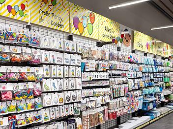 Ein gut sortiertes Ladenregal voller Partyartikel wie Ballons und Dekorationen. Die Beschriftung oben lautet: "Gro&szlig;e Party, kleine Kosten" und "Alles f&uuml;r deine Party".