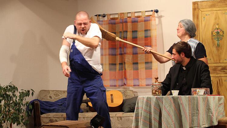 Mit handfesten Argumenten versucht Pfarrhaushälterin Hermine (Martina Lutter) den Mesner Johannes Höll (Jens Müller) zur Arbeit zu bewegen. Pfarrer Alfons (Philipp Gernlein) schaut dem Treiben zu.