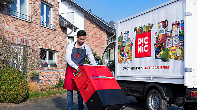 Ein Mann in einer Schürze liefert mit einem roten Handwagen Lebensmittel aus einem Picnic-Lieferwagen vor einem Backsteinhaus.