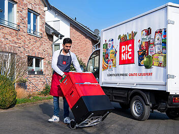Ein Mann in einer Schürze liefert mit einem roten Handwagen Lebensmittel aus einem Picnic-Lieferwagen vor einem Backsteinhaus.