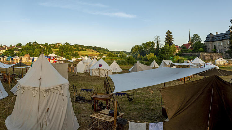 Ein mittelalterliches Lager mit zahlreichen weißen Zelten auf einer Wiese. Im Hintergrund sind Gebäude und Bäume unter blauem Himmel zu sehen. Es vermittelt eine historische Atmosphäre.