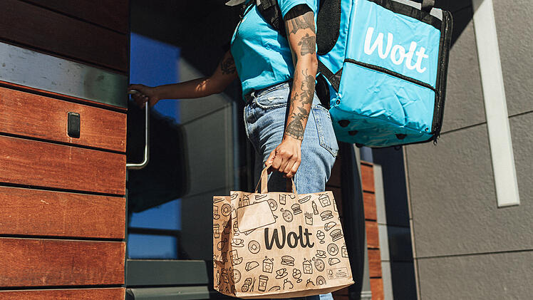 Ein Wolt-Lieferfahrer tr&auml;gt eine blaue Lieferbox und eine braune Papiert&uuml;te mit dem Wolt-Logo zu einer T&uuml;r. Die Szene zeigt den Lieferdienst in Aktion.
