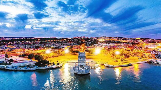 Lisbon, Portugal. Aerial view of Belem Tower in Lisbon, Portugal during the sunset. Illuminated buildings with cloudy sky and ocean. Lissabon, Portugal. Luftaufnahme des Belem-Turms in Lissabon, Portugal, während des Sonnenuntergangs. Beleuchtete Gebäude mit bewölktem Himmel und Meer. Luftaufnahme des Belem-Turms in Lissabon, Portugal, während des Sonnenuntergangs