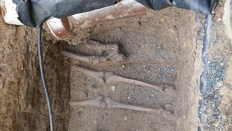 Das Bild zeigt arch&auml;ologische Ausgrabungen mit menschlichen Knochen, die in Erde eingebettet sind. Daneben sind ein Rohr und ein Kabel sichtbar.