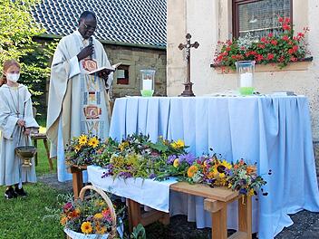 Abb&eacute; Pierre spricht das Segensgebet &uuml;ber die farbenpr&auml;chtigen Kr&auml;uterb&uuml;schel und besprengt sie mit Weihwasser. Links im Bild Ministrantin Marlene Pfeuffer, dahinter das &bdquo;H&ouml;reder&ldquo; Pfarrhaus mit einladendem Blumenschmuck.