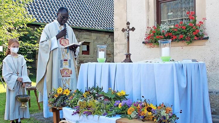 Abbé Pierre spricht das Segensgebet über die farbenprächtigen Kräuterbüschel und besprengt sie mit Weihwasser. Links im Bild Ministrantin Marlene Pfeuffer, dahinter das „Höreder“ Pfarrhaus mit einladendem Blumenschmuck.