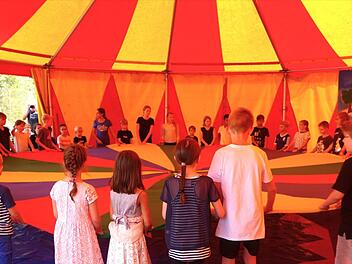 Ein buntes Zelt mit spielenden Kindern unter einem farbenfrohen Tuch. Der Ort wirkt einladend und fröhlich, mit Raum für Lernen und Gemeinschaft.