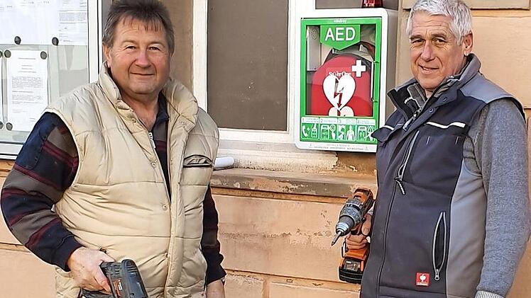 Die Vorsitzenden Rainer Schüll von der Feuerwehr (links) und Reinhold Klein vom Sportverein (rechts) brachten den Defibrillator am Haus der Vereine in Gemeinfeld an. Foto: privat