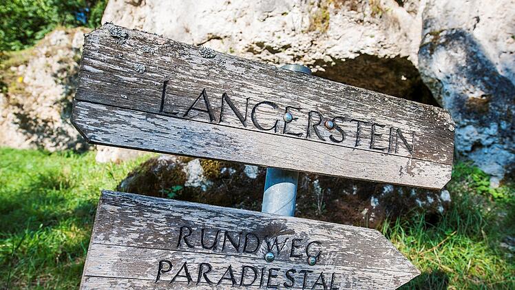 Ein verwittertes Holzschild zeigt den Weg zu "Langerstein" und "Rundweg Paradiestal", vor einem Felsen im Gr&uuml;nen in der Fr&auml;nkischen Schweiz.