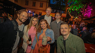 Eine Gruppe von Menschen posiert fröhlich bei der Bamberger Sandkerwa. Hintergrund: festliche Lichter, ein Baum und festliche Stimmung.