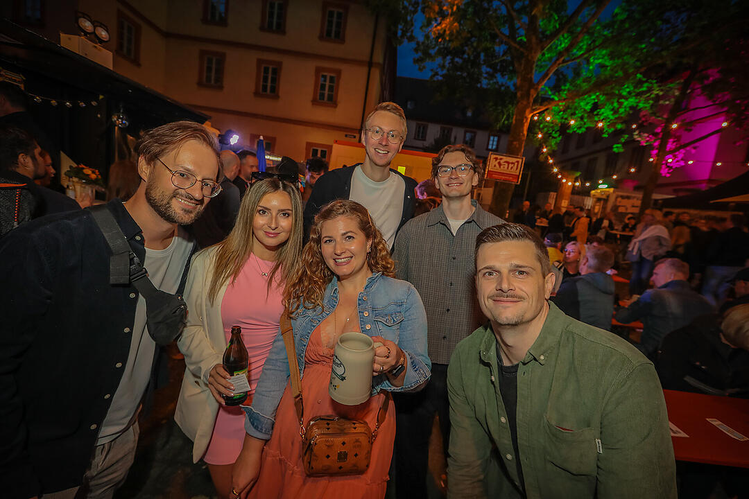Eine Gruppe von Menschen posiert fröhlich bei der Bamberger Sandkerwa. Hintergrund: festliche Lichter, ein Baum und festliche Stimmung.