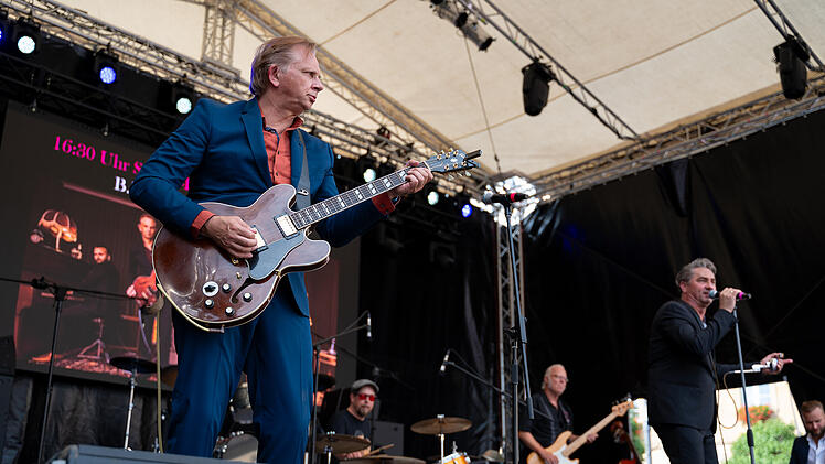 Musiker auf einer B&uuml;hne beim Blues- und Jazzfestival in Bamberg. Ein Gitarrist in einem blauen Anzug spielt E-Gitarre, w&auml;hrend ein S&auml;nger ins Mikrofon singt. Hinweis auf eine Show um 16:30 Uhr.
