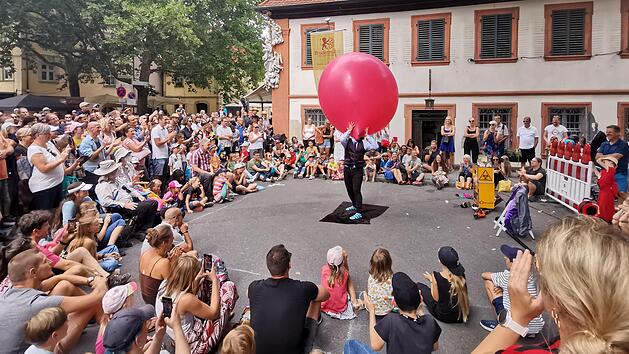 Ein K&uuml;nstler in der Mitte einer Menschenmenge h&auml;lt einen gro&szlig;en roten Ballon. Die Zuschauer, darunter viele Familien, sind begeistert und schauen gespannt zu. Hintergrund: ein altes Geb&auml;ude.