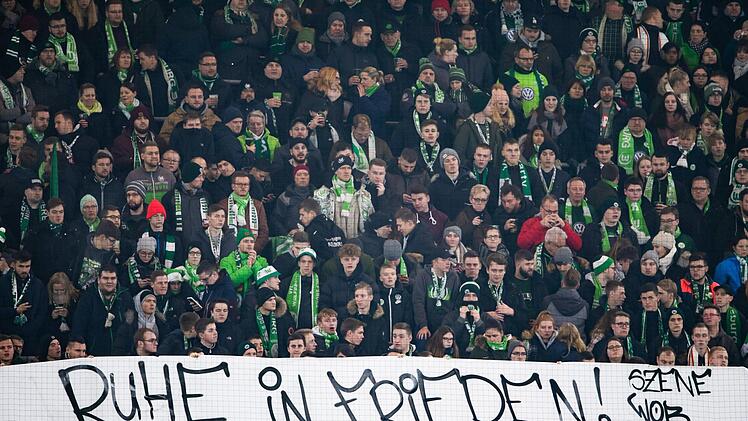 Fans vom VfL Wolfsburg halten ein Plakat mit der Aufschrift "Ruhe in Frieden!". Auf dem Weg zum Fu&szlig;ballspiel sein ein Fan des FC Schalke 04 in der N&auml;he des Stadions zusammengebrochen und verstarb sp&auml;ter, sagte ein Polizeisprecher. Foto: Swen Pf&ouml;rtner/dpa