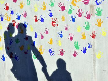 Ein Mann mit einem Kind auf dem Arm und einem an der Hand wirft einen Schatten auf eine mit bunten Handabdrücken bemalte Wand einer Kindertagesstätte.  Foto: Peter Kneffel/dpa