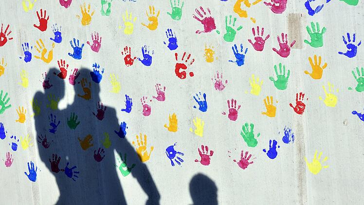 Ein Mann mit einem Kind auf dem Arm und einem an der Hand wirft einen Schatten auf eine mit bunten Handabdrücken bemalte Wand einer Kindertagesstätte.  Foto: Peter Kneffel/dpa