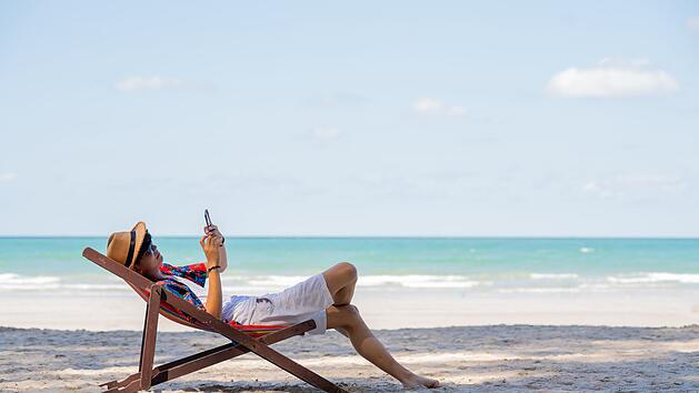 Junger asiatischer Mann, der sich auf einer Sonnenliege an einem tropischen Strand ausruht. Fröhlicher Mann sitzt auf einem Strandstuhl am Meer und nutzt sein Smartphone für Selfies oder Videoanrufe. Attraktiver Mann genießt den Strand und das Leben im Freien während seines Sommerurlaubs. Young Asian man resting on sunbed on tropical beach. Happy guy sit on beach chair by the sea using smartphone for selfie or video call. Handsome male enjoy beach outdoor lifestyle on summer vacation