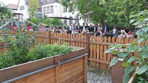 Schüler und Schülerinnen, die Lehrerschaft und die geladenen Gäste bestaunen den neu angelegten Schulgarten der Pestalozzi Grundschule.