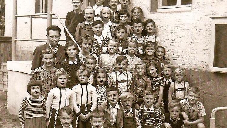 Diese Aufnahme zeigt den Schuljahrgang 1955/56. Unterrichtet wurden die Kinder von Lehrer Franz Buschbacher (3. Reihe, links). Sein gleichnamiger Sohn, der Co-Autor des Buches, ist in der 2. Reihe als Zweiter von rechts zu sehen, dessen Zwillingsschwestern stehen in der dritten Reihe und tragen gepunktete Kleider.