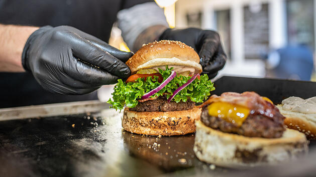 Ein Burger wird von einer Person mit schwarzen Handschuhen zubereitet. Frische Zutaten wie Salat, rote Zwiebeln und ein Patty sind zu sehen. Im Hintergrund verschwommenes Innenraum-Ambiente.