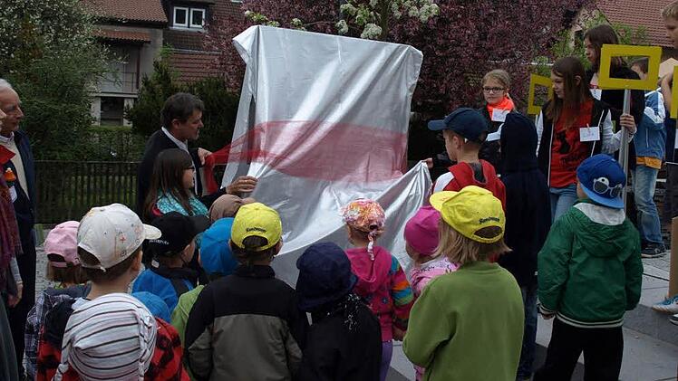Bürgermeister Gerhard Wunder enthüllt zusammen mit den Kindern den Bilderrahmen am Rathausplatz mit Blick auf die katholische Pfarrkirche.