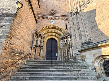 Historisches Kirchenportal mit verzierten Statuen flankiert einen schmalen Eingang. Steinstufen f&uuml;hren hinauf. Architektonische Details reflektieren das Licht; m&ouml;glicherweise Bamberger Dom.