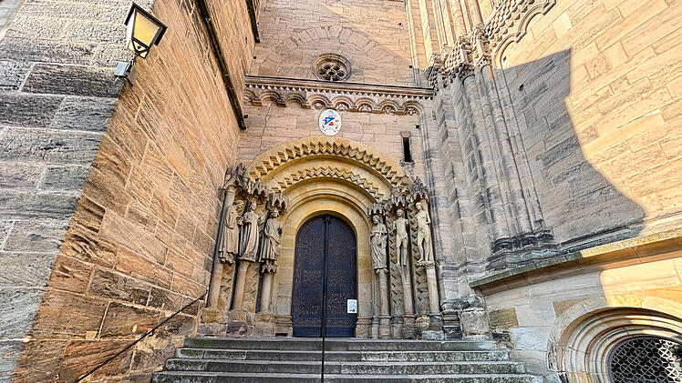 Historisches Kirchenportal mit verzierten Statuen flankiert einen schmalen Eingang. Steinstufen f&uuml;hren hinauf. Architektonische Details reflektieren das Licht; m&ouml;glicherweise Bamberger Dom.