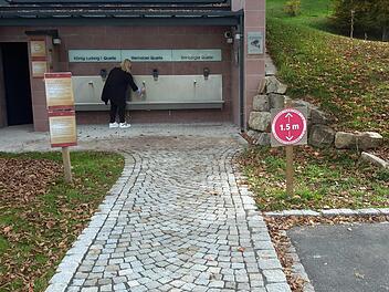 Kurdirektorin Andrea Schallenkammer (hinten) und ihr Stellvertreter Titus Tesar (rechts) demonstrieren den Ablauf. Nur jeweils eine Person darf die Zapfstelle mit Mund-Nasen-Schutz betreten, draußen vor der Anlage ist ein Mindestabstand von 1,5 Metern zu wahren. Foto: Rolf Pralle