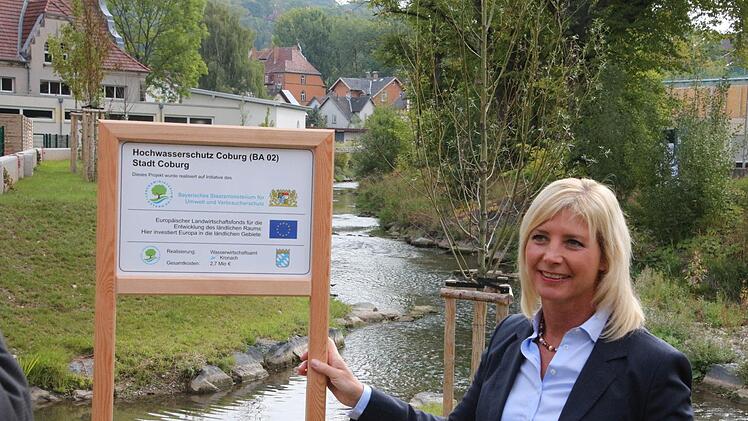 Frau vor Bachlandschaft: Bayerns Umweltministerin Ulrike Scharf vor dem neuen Zusammenfluss von Rottenbach und Lauter. Die Tafel informiert darüber, was dieser Abschnitt des Hochwasserschutzes gekostet hat. Foto: Simone Bastian