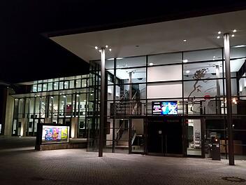 Bamberg: ETA Hoffmann Theater lädt zu Blick hinter die Kulissen ein - Infos und Anmeldung Das Bild zeigt ein modern beleuchtetes Gebäude bei Nacht mit Glasfassade. Links sind bunte Plakate, rechts ein großes Porträt an der Wand. Eine digitale Anzeigetafel ist sichtbar.