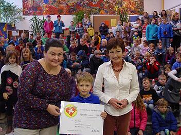 Phil Parzer (Mitte) übergibt den Spenden-Scheck an Daniela Schad (links). Mit ihnen freuen sich Schulleiterin Barbara Buz (rechts) und die vielen Kinder im Hintergrund. Foto: Brigitta Kempe-Wolf