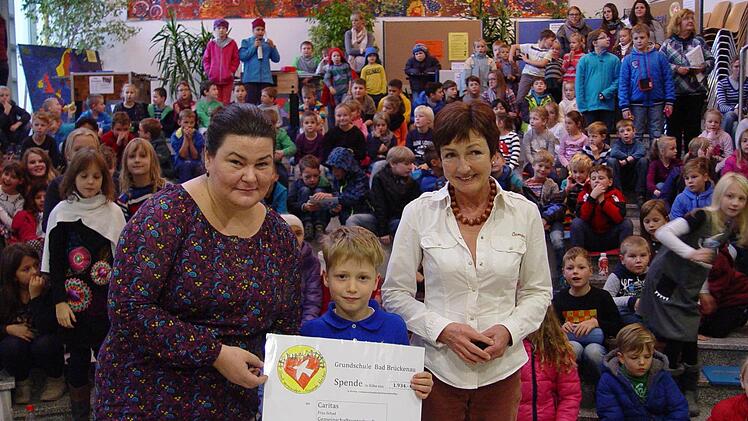Phil Parzer (Mitte) übergibt den Spenden-Scheck an Daniela Schad (links). Mit ihnen freuen sich Schulleiterin Barbara Buz (rechts) und die vielen Kinder im Hintergrund. Foto: Brigitta Kempe-Wolf