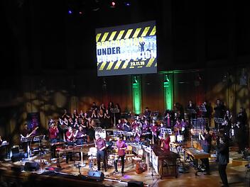 Ganz schön eng wurde es auf der Bühne im Max-Littmann-Saal bei Thomas Friedrichs Großveranstaltung in der "7. Nacht der Percussion": Hinten links der Hausener Chor Chorisma mit seiner Leiterin Antje Kopp, hinten rechts die Bläserband, vorne rechts das Cello von Claudia Dunkelberg, vorne links E-Gitarrist und Keyboarderin, im Zentrum Kisspercussiva, deren Leiter Thomas Friedrich von rechts begeistert applaudiert. Gerhild Ahnert