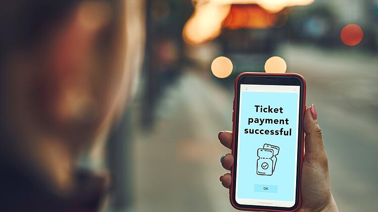 Frau kauft vor der Fahrt an der Bushaltestelle online ein Ticket. Person bezahlt das Ticket mit einer mobilen Zahlungs-App. Nutzung von Technologie unterwegs. Bestellbestätigung auf dem Handybildschirm. Konto in der App aufladen. Busbeleuchtung im Hintergrund. Woman buying ticket online at bus stop before ride. Person paying for ticket using mobile payment app. Using technology on the go. Order confirmation on mobile screen. Top up account in app. Bus lights in background