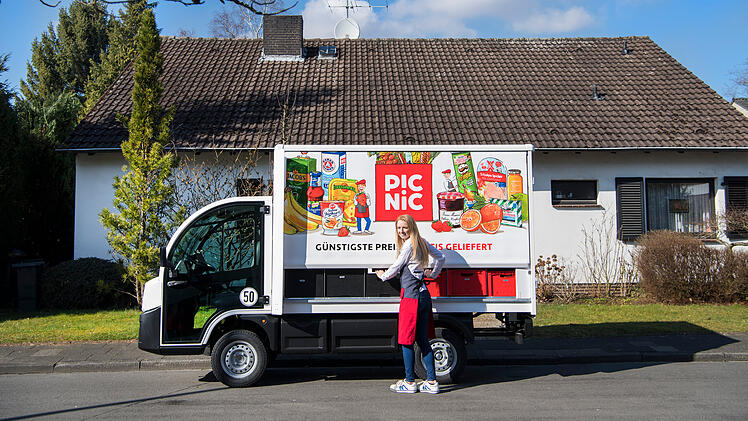 Ein Lieferwagen mit "Picnic"-Logo steht vor einem Haus. Eine Person in Schürze steht daneben. Der Himmel ist blau, teils bewölkt, und um das Haus sind Bäume und Büsche zu sehen.