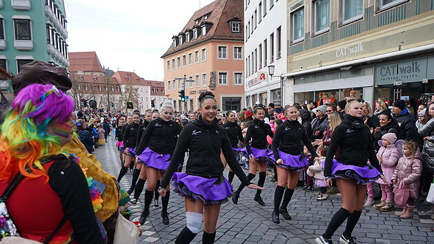 Eine fr&ouml;hliche Faschingsparade zieht durch die belebte Stra&szlig;e. T&auml;nzerinnen in lila R&ouml;cken f&uuml;hren auf, w&auml;hrend bunt gekleidete Zuschauer applaudieren und zuschauen.