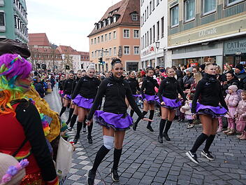 Eine fröhliche Faschingsparade zieht durch die belebte Straße. Tänzerinnen in lila Röcken führen auf, während bunt gekleidete Zuschauer applaudieren und zuschauen.