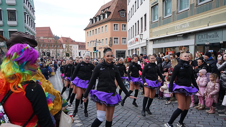 Eine fröhliche Faschingsparade zieht durch die belebte Straße. Tänzerinnen in lila Röcken führen auf, während bunt gekleidete Zuschauer applaudieren und zuschauen.