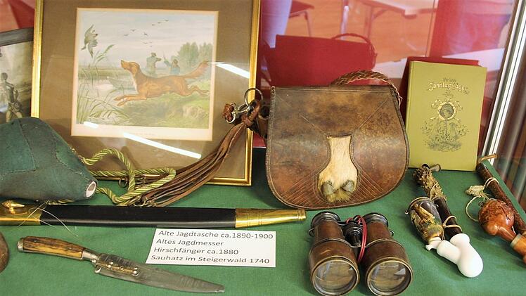Eine historische Jagdtasche (Ende des 19. Jahrhunderts) liegt neben einem antiken Fernglas, Jagdmessern und reich verzierten Jäger-Pfeifen. Fuchs