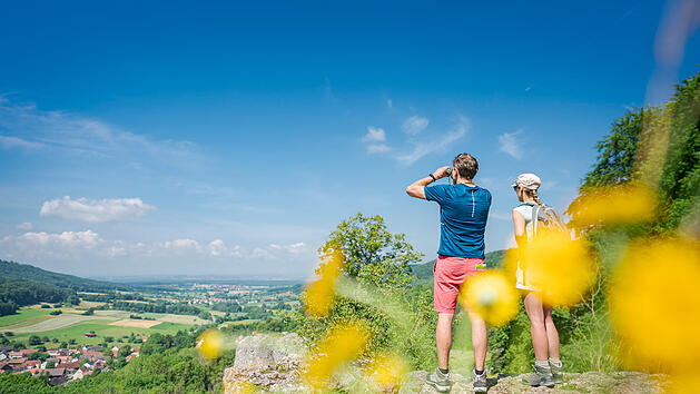 Ein Paar steht auf einer Klippe in der "Fr&auml;nkischen Toskana", genie&szlig;t die Aussicht auf gr&uuml;ne Felder und W&auml;lder. Im Vordergrund gelbe Blumen, der Himmel ist klar und blau.