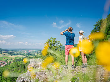 Ein Paar steht auf einer Klippe in der "Fr&auml;nkischen Toskana", genie&szlig;t die Aussicht auf gr&uuml;ne Felder und W&auml;lder. Im Vordergrund gelbe Blumen, der Himmel ist klar und blau.