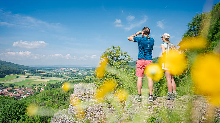 Ein Paar steht auf einer Klippe in der "Fr&auml;nkischen Toskana", genie&szlig;t die Aussicht auf gr&uuml;ne Felder und W&auml;lder. Im Vordergrund gelbe Blumen, der Himmel ist klar und blau.