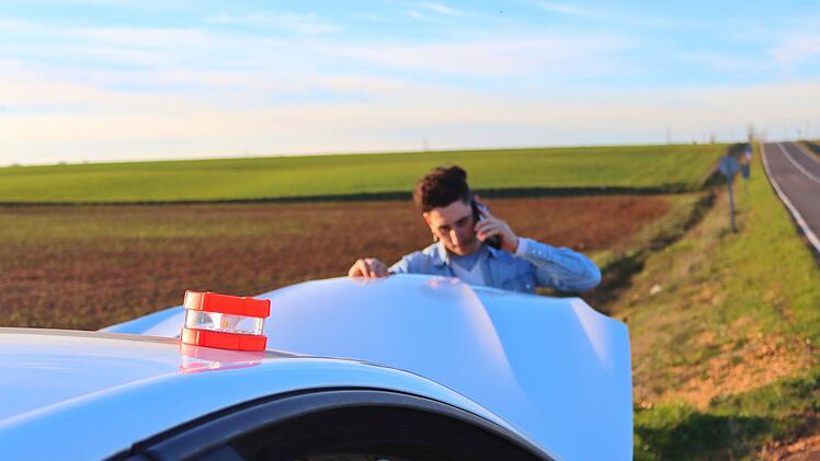 Ab dem 1. Januar 2026 modernisiert Spanien die Sicherheitsvorkehrungen im Straßenverkehr. Das traditionelle Warndreieck hat ausgedient und wird durch die innovative V16-Warnleuchte ersetzt. Kaukasischer Mann telefoniert und schaut auf den Motor mit der Notfallleuchte V16, tagsüber. Konzept eines Problems auf der Straße  von Sergio Caucasian man calling on the phone looking at the engine with the emergency light V16, daytime. Concept of problem on road