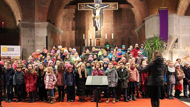 Kinderchor probt in einer Kirche für ein Weihnachtskonzert. Dirigentin steht vorne, viele Kinder bunt gekleidet. Hintergrund zeigt Kruzifix und Kerzen.